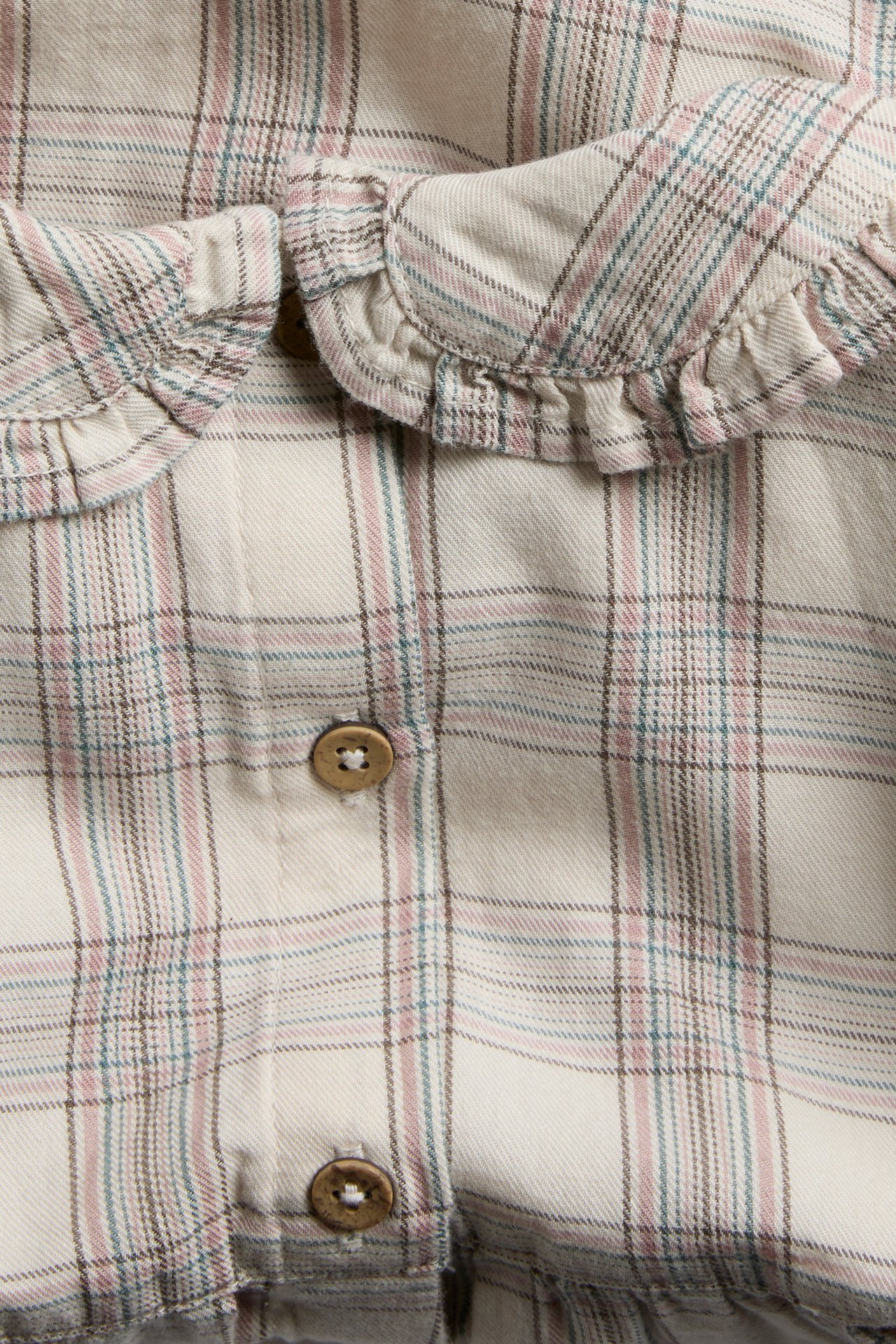 Close-up of a beige plaid shirt with pink, blue, and brown lines, featuring a ruffled collar and wooden buttons.