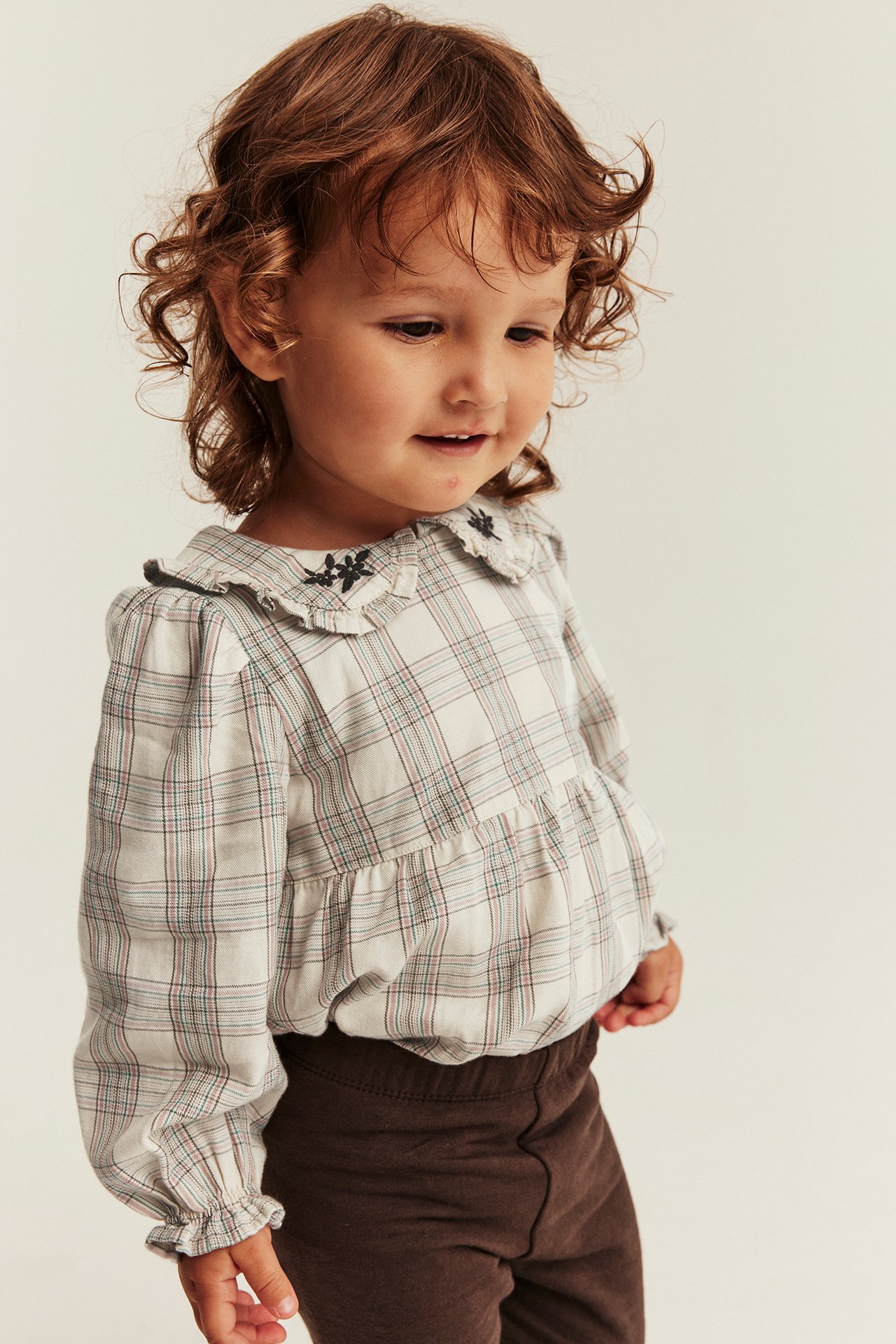 Front view of a kid wearing a white plaid long-sleeved shirt with embroidered ruffled collar and brown leggings.
