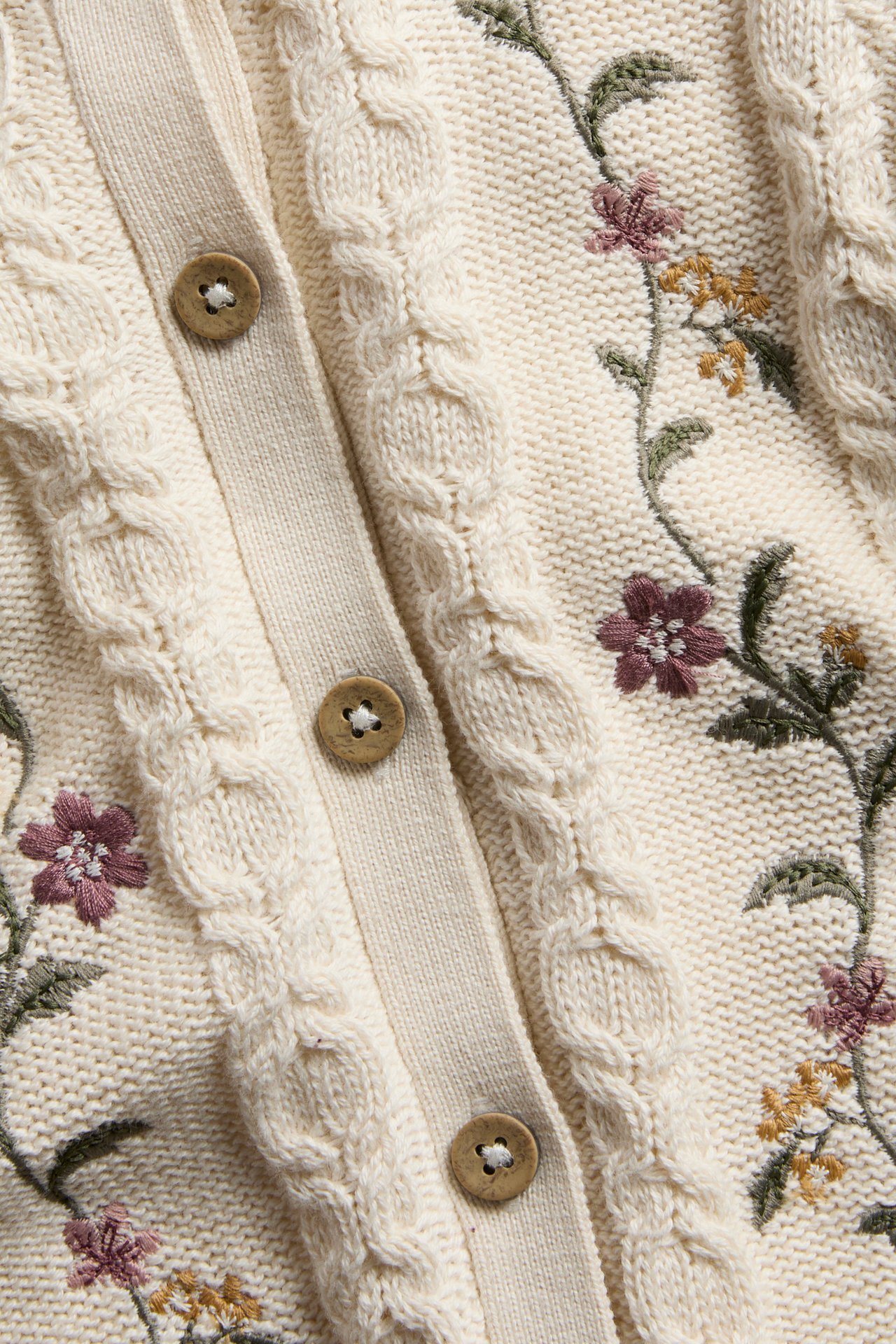 Nahaufnahme einer cremefarbenen Zopfmuster-Strickjacke mit Blumenstickerei und Holzknöpfen.