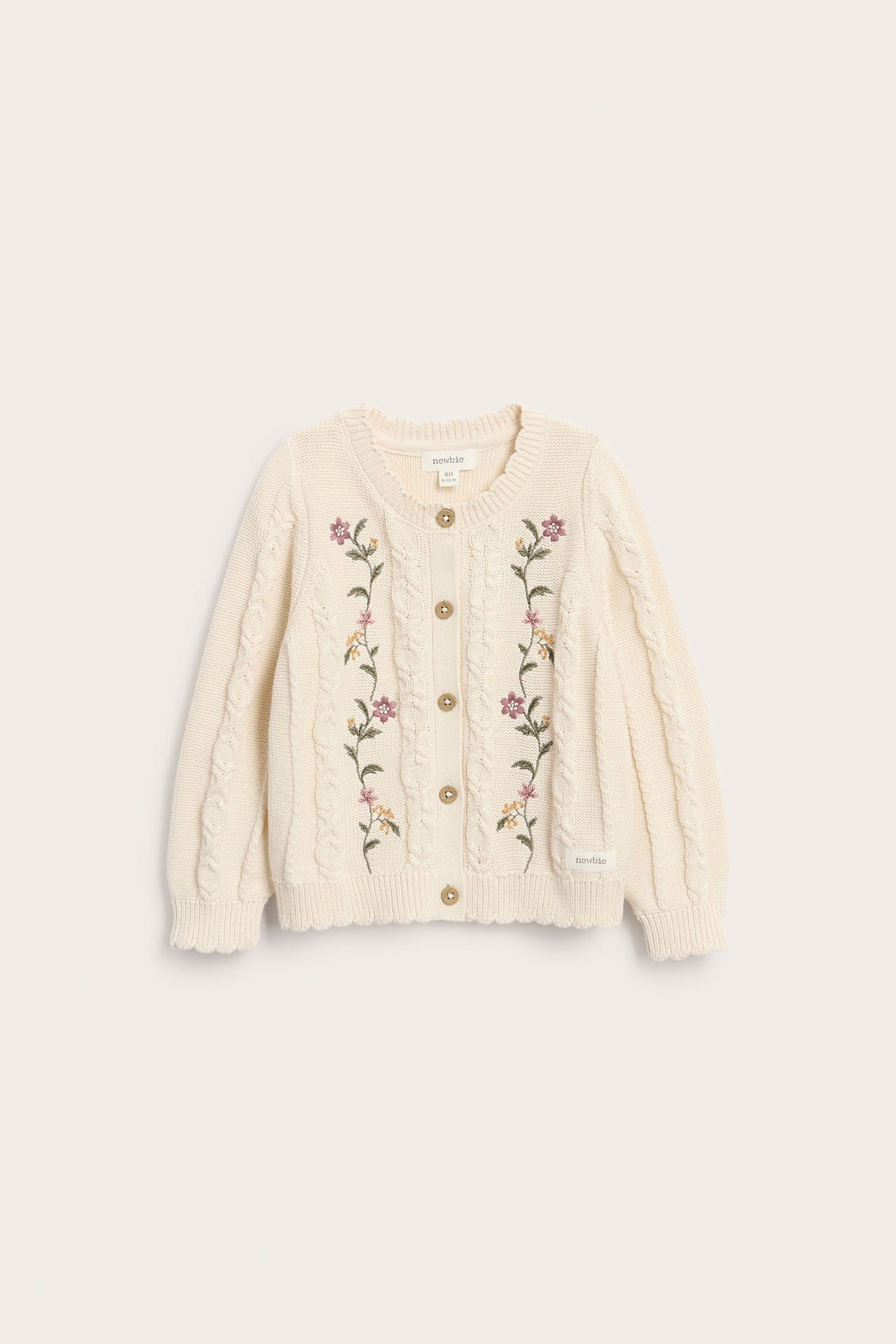 Vorderansicht einer cremefarbenen Kinder-Strickjacke mit Zopfmuster, Blumenstickerei und Holzknöpfen.