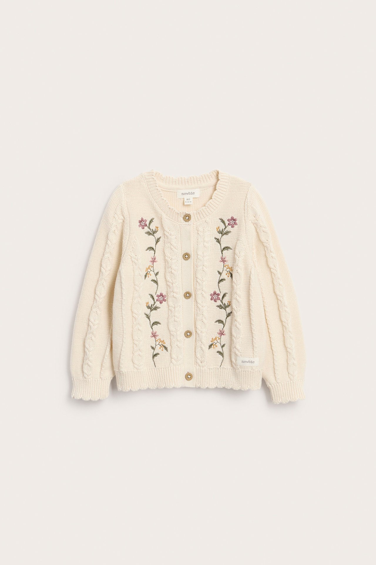 Vorderansicht einer cremefarbenen Kinder-Strickjacke mit Zopfmuster, Blumenstickerei und Holzknöpfen.