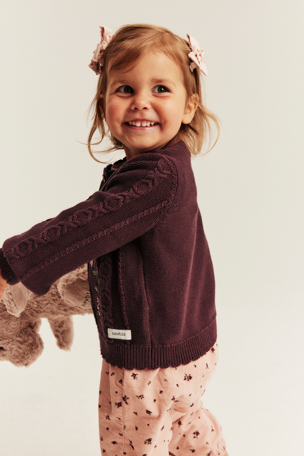 Seitenansicht eines Kindes in einer dunkel pflaumenfarbenen Zopfstrickjacke und rosa geblümten Hose, hält ein beiges Spielzeug.