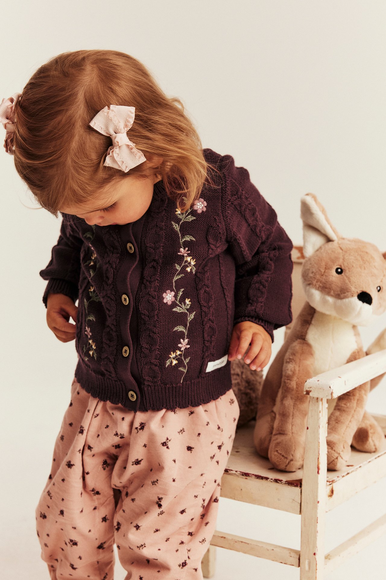 Vorderansicht: Kind in dunkelvioletter bestickter Zopfstrickjacke, rosa geblümten Hosen und rosa Haarschleifen.