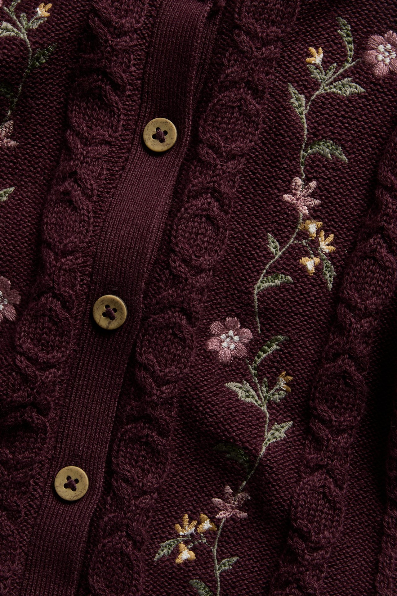 Nahaufnahme eines burgunderfarbenen Strickcardigans mit Zopfmuster, Blumenstickerei und braunen Knöpfen.