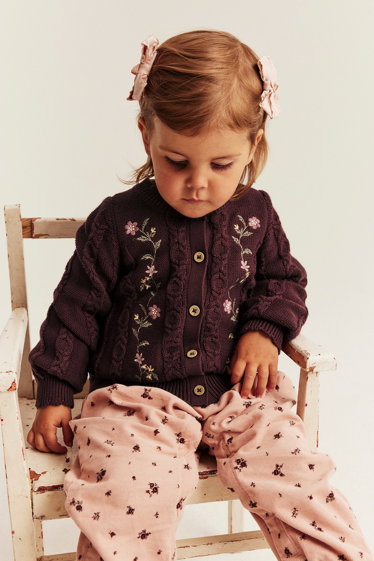 Kind in dunkellila Zopfstrickjacke mit Blumenstickerei, rosa Blumenhose und rosa Schleifen.