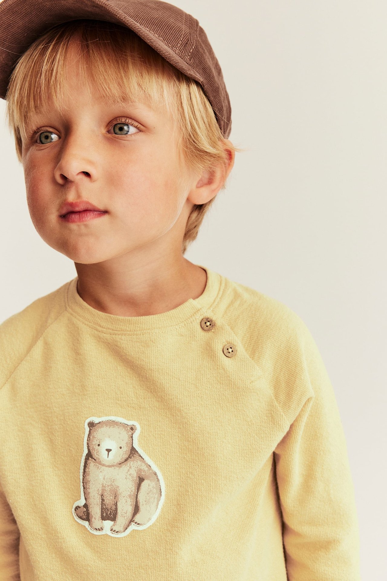 Nahaufnahme von Kind in gelbem Sweatshirt mit Bärendruck und brauner Mütze.