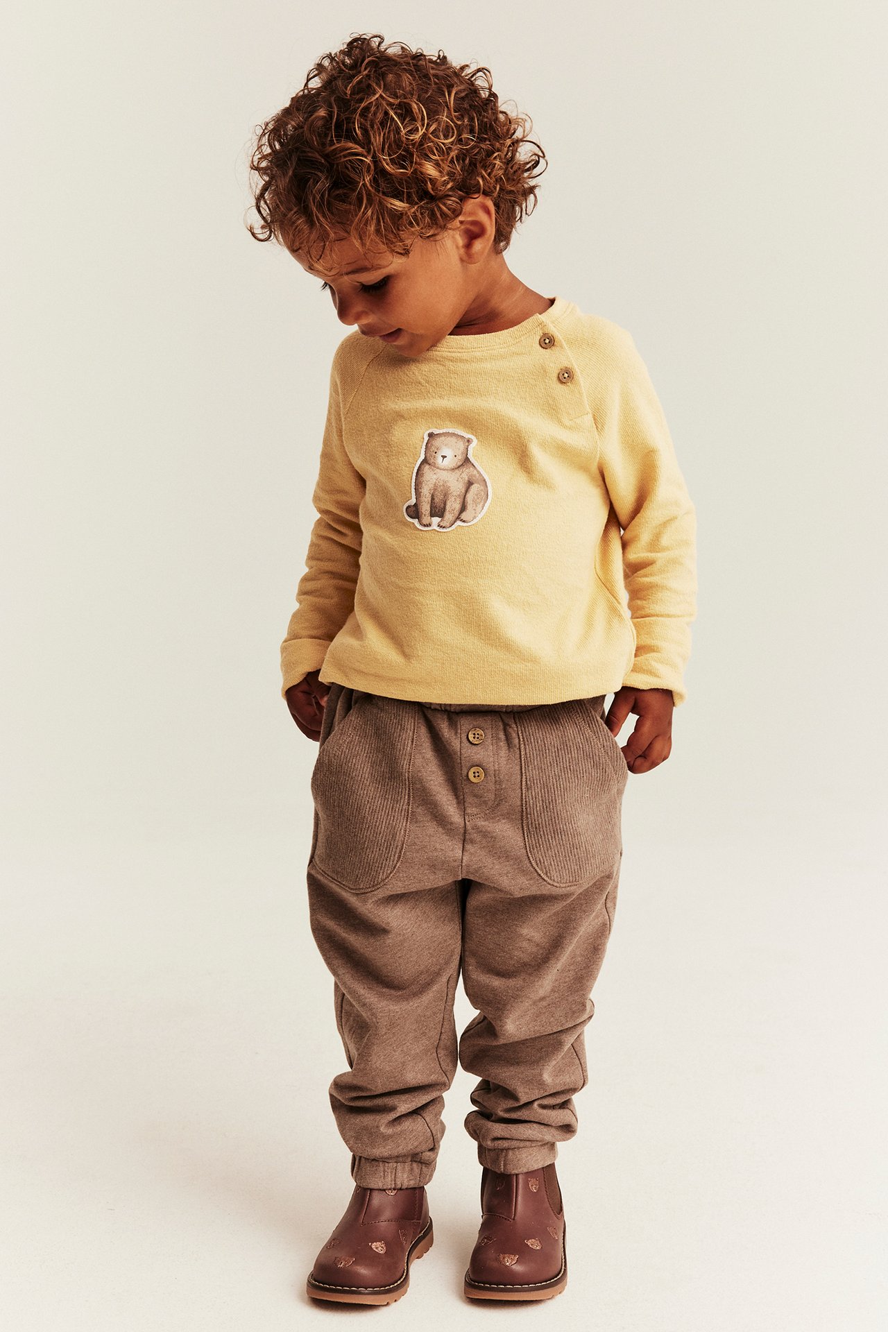 Vorderansicht eines Kindes in gelbem Langarmshirt mit Bären-Patch, brauner Cordhose und braunen Stiefeln.