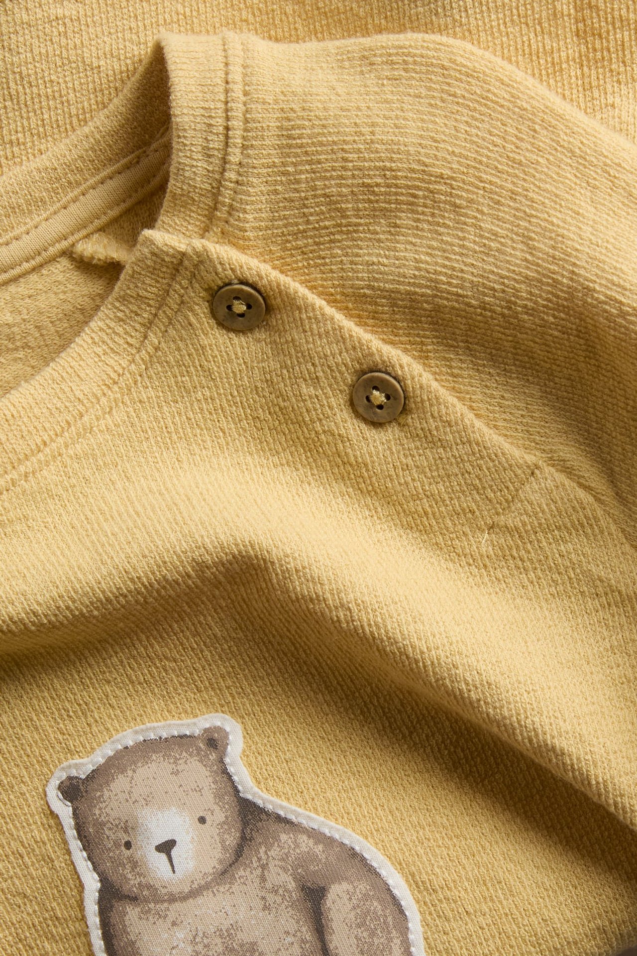 Close-up of a yellow ribbed baby top with shoulder snap buttons and a bear appliqué.