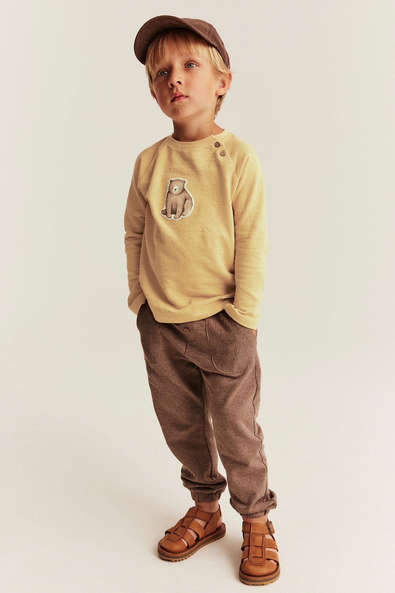 Front view of a kid in a yellow bear patch top, brown ribbed joggers, brown sandals, and a brown cap.