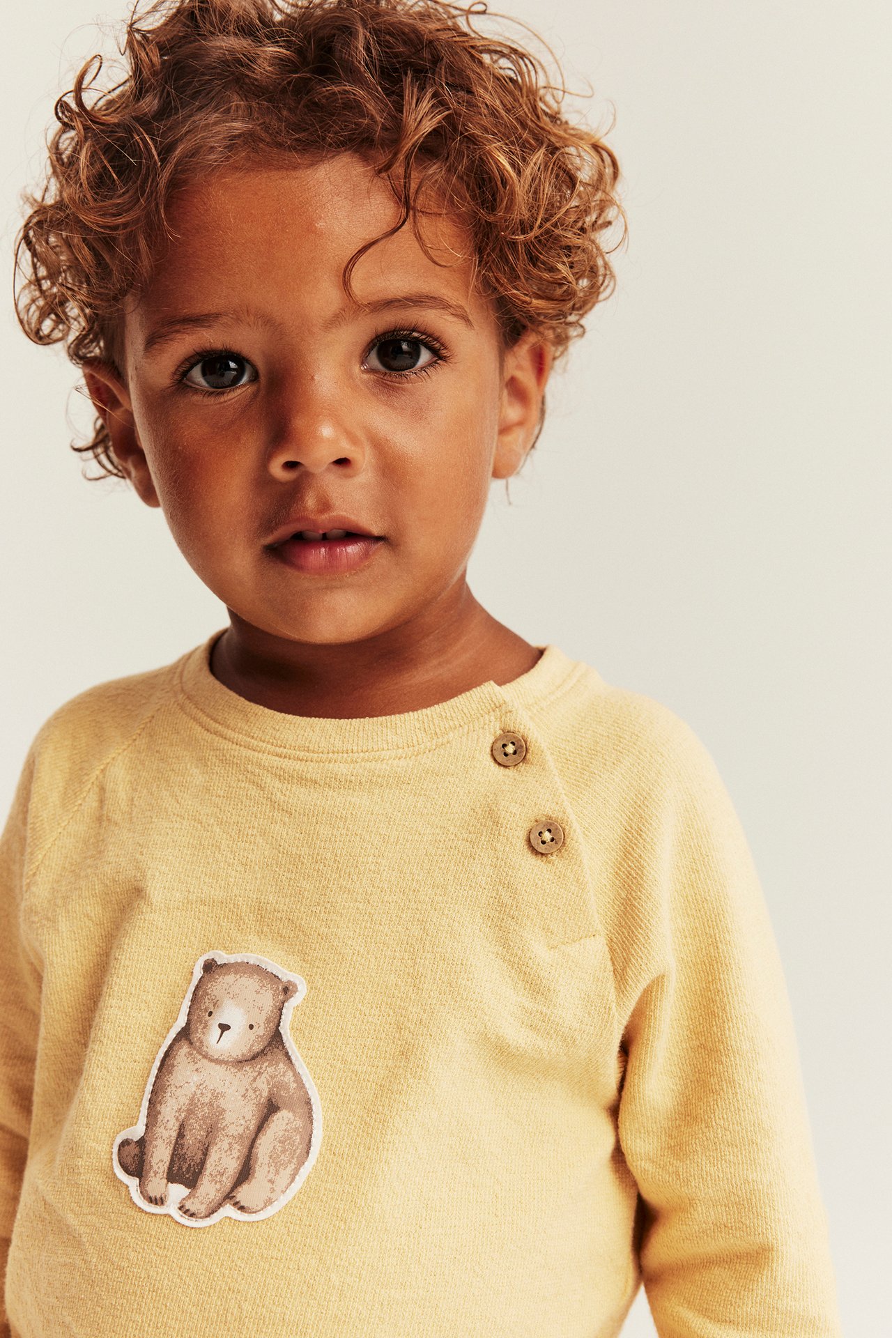 Close-up front view of a kid wearing a yellow long-sleeved top with shoulder buttons and a bear patch.