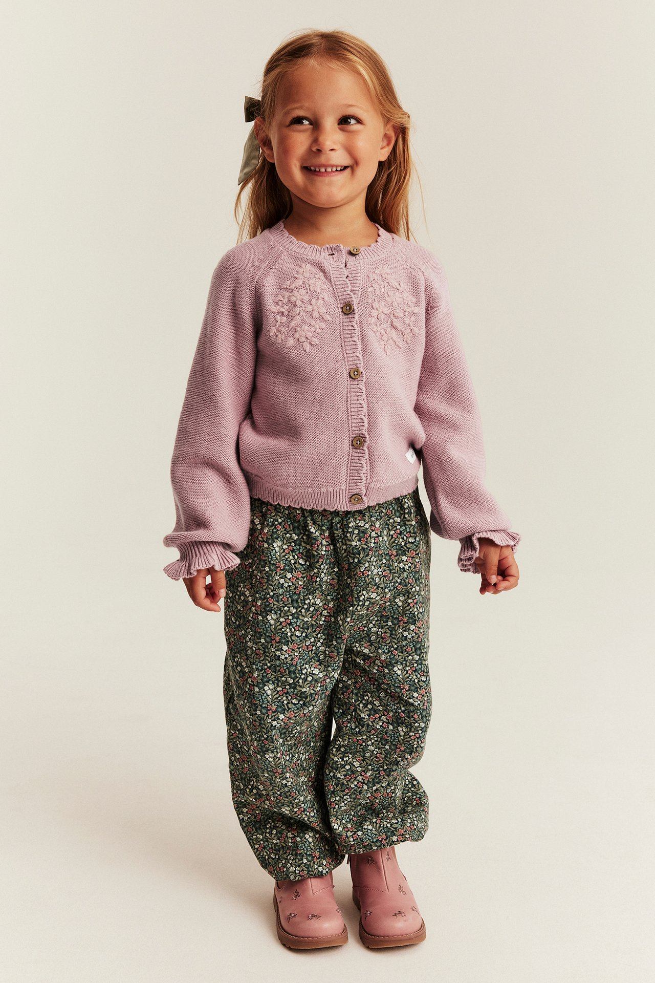 Vorderansicht eines Kindes in hellviolettem besticktem Cardigan, dunkelgrüner Blumenhose und rosa Stiefeln.