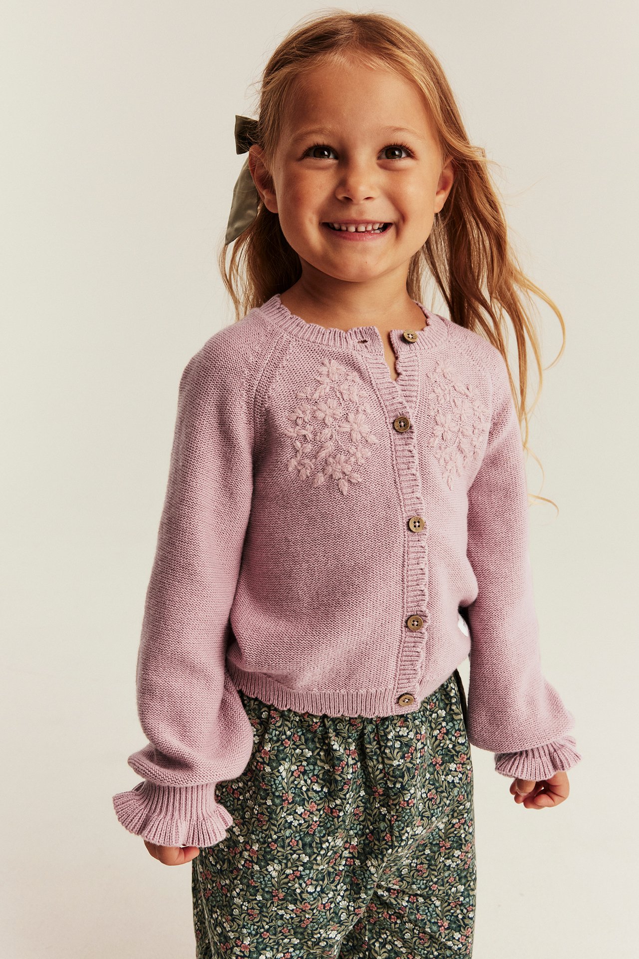 Vorderansicht eines Kindes in rosa Strickjacke mit Blumenstickerei und gemusterter Hose.