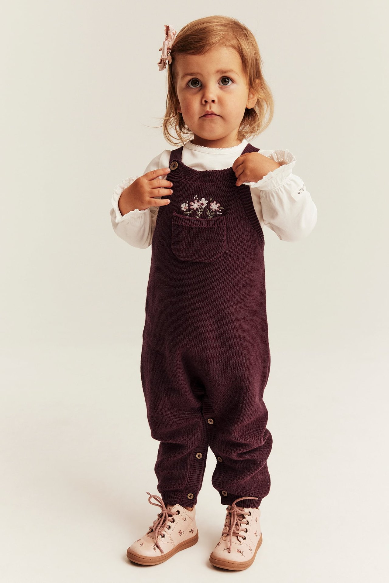 Vorderansicht eines Babys in einem burgunderroten Strick-Overall mit bestickter Blumentasche, weißem Top, rosa Stiefeln und Haarspange.