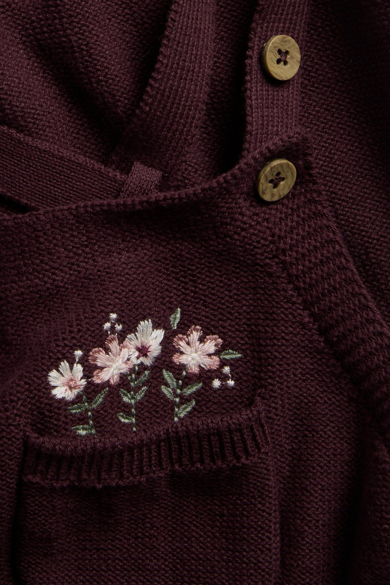 Nahaufnahme eines burgunderfarbenen Strickkleidungsstücks mit Blumenstickerei und zwei Holzknöpfen.