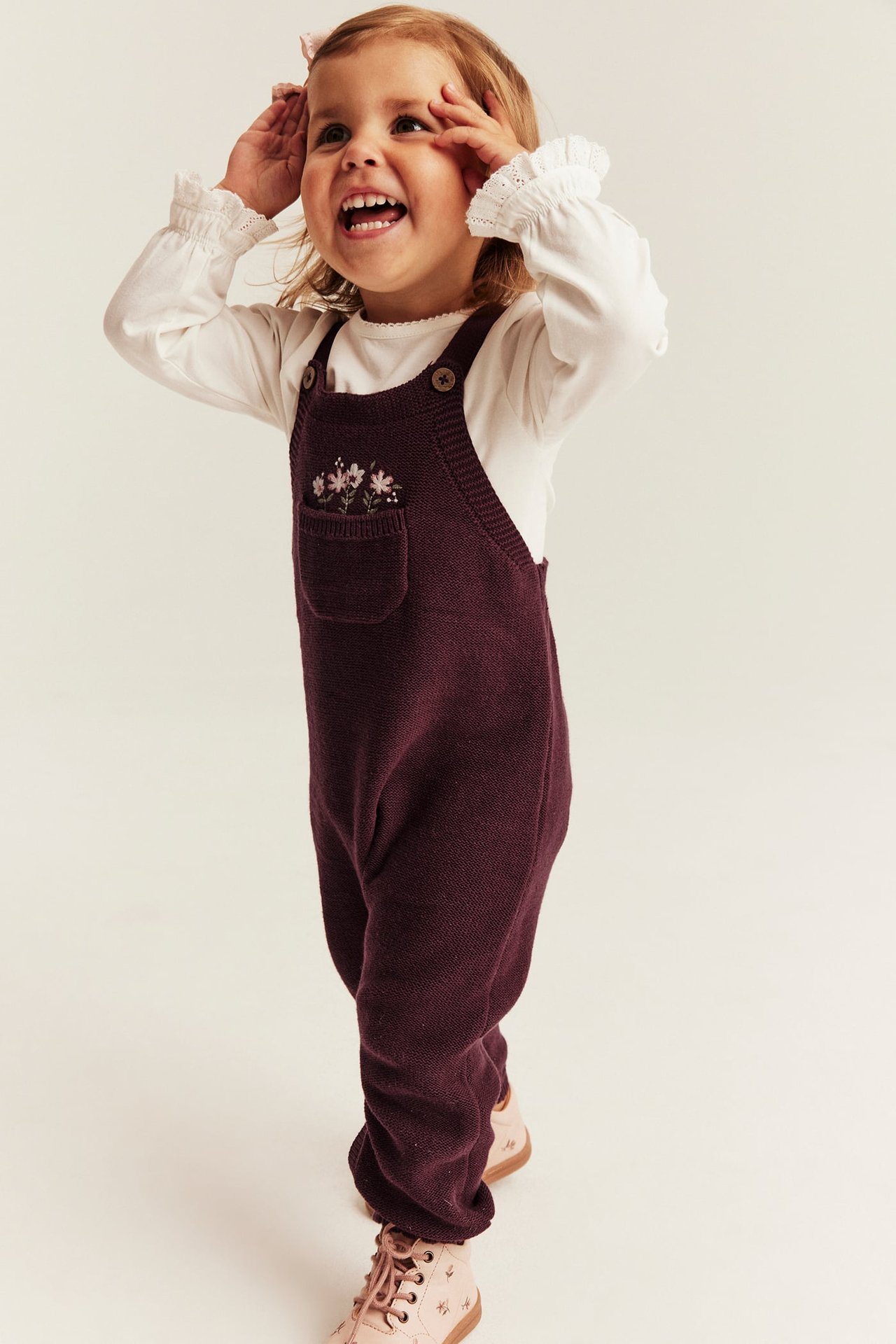 Vorderansicht: Burgunderroter Strick-Overall mit Blumenstickerei, weißes Langarmshirt und rosa Stiefel.