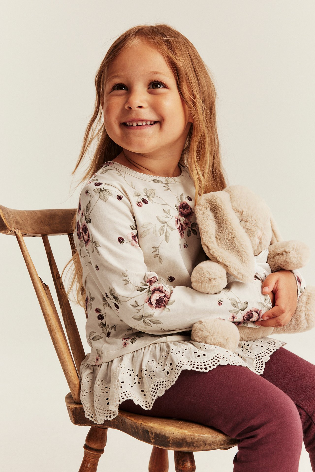 Kinder cremefarbenes Langarmshirt mit Blumenmuster, Spitze und bordeauxroten Leggings, hält einen beigefarbenen Hasen.