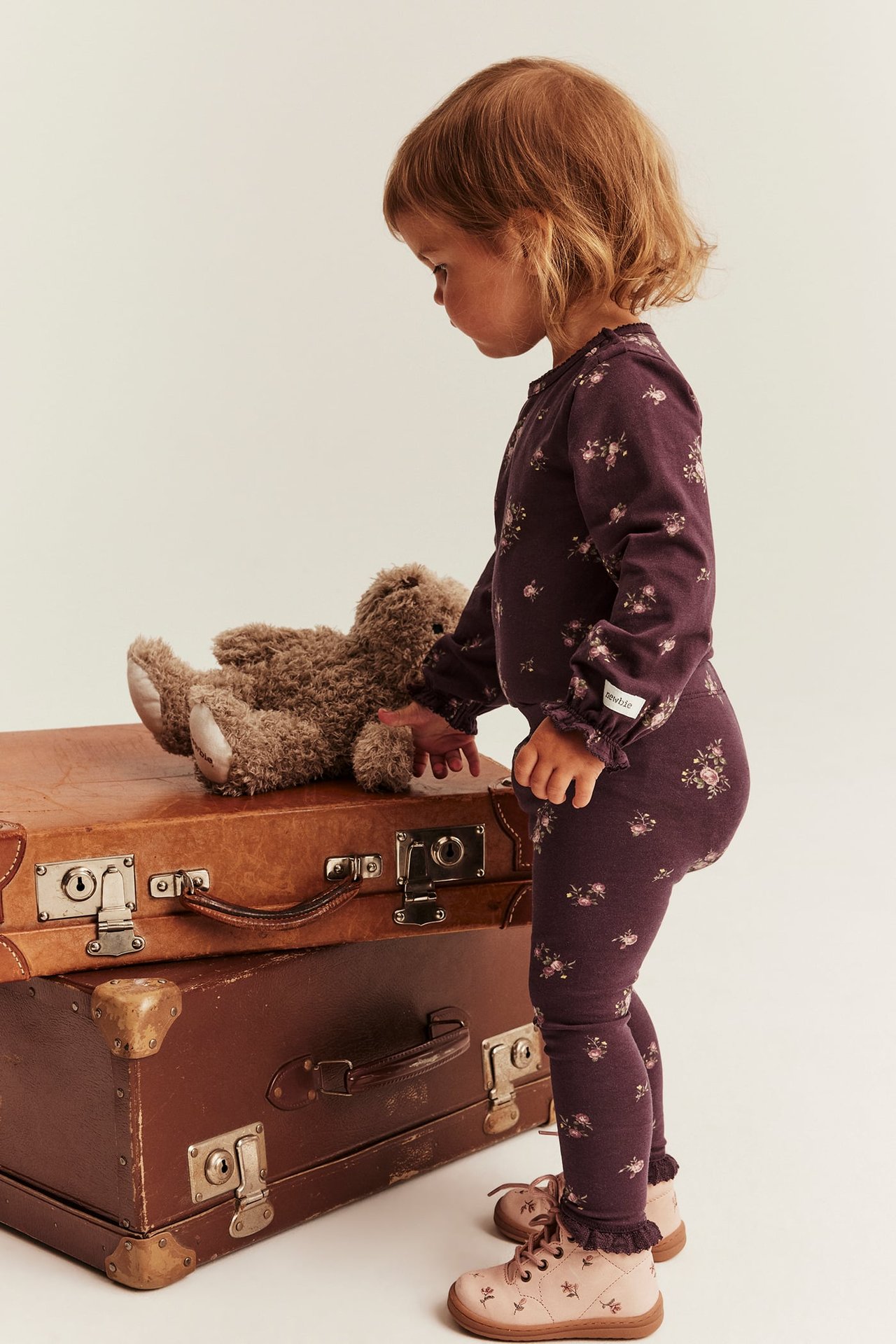 Seitenansicht: Kind in lila Blumen-Set, rosa Schuhen, berührt Teddybär auf Koffern.