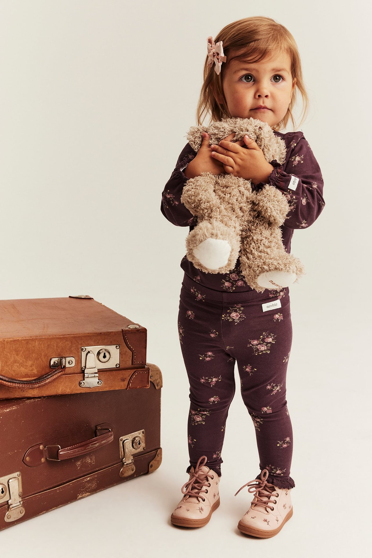 Vorderansicht eines Babys in dunkelviolettem Blumen-Langarmshirt, passenden Leggings und rosa Stiefeln, hält einen Teddy.