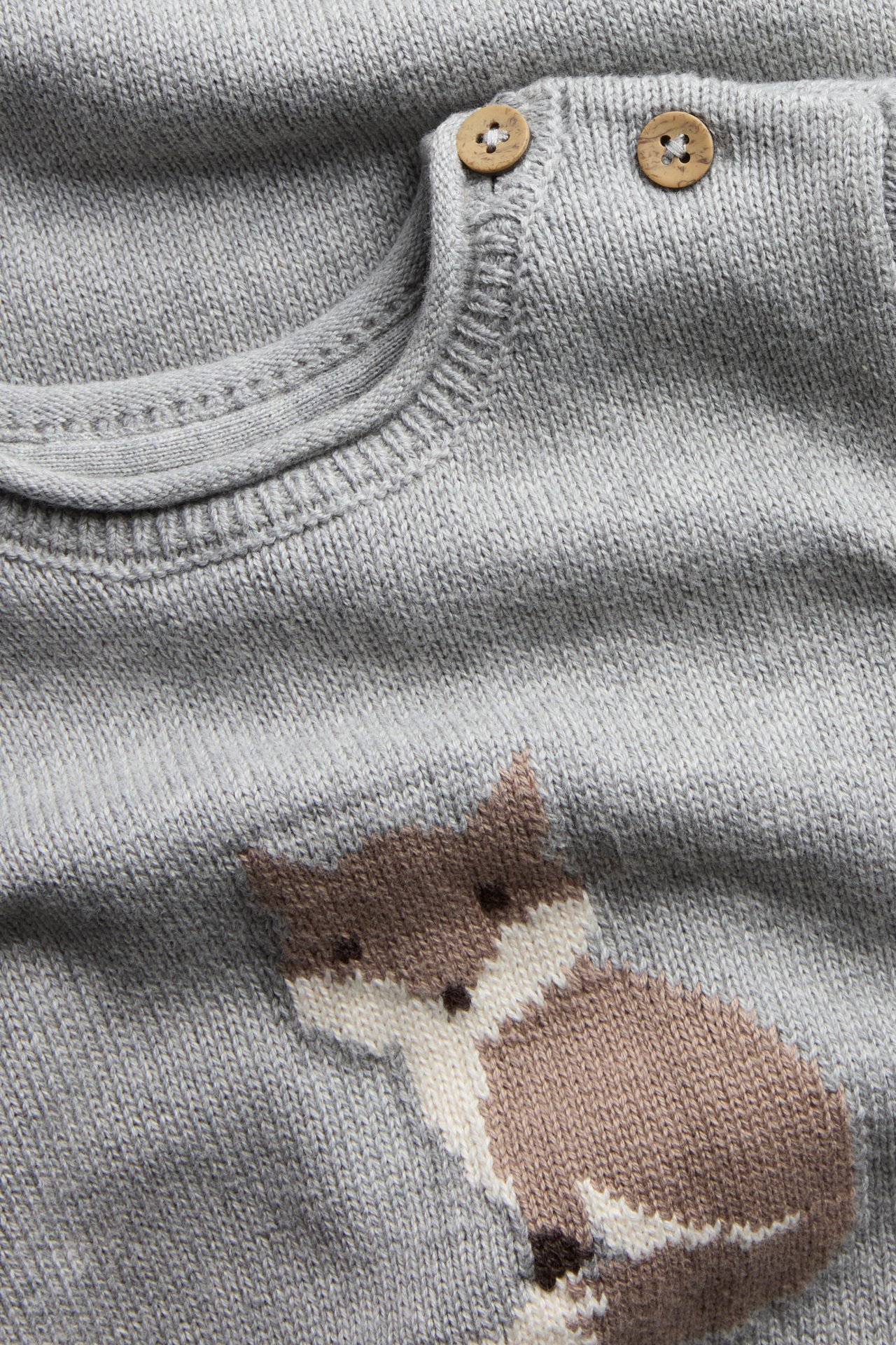 Nahaufnahme eines grauen Baby-Strickpullovers mit braun-weißem Fuchsmotiv und zwei Holzknöpfen an der Schulter.