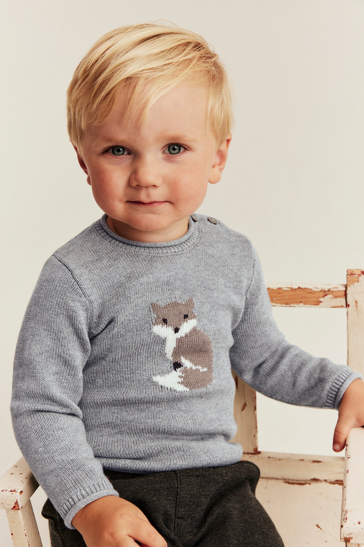 Kleiner Junge in einem hellgrauen Strickpullover mit Fuchsmotiv und dunkelgrauer Hose, Vorderansicht.