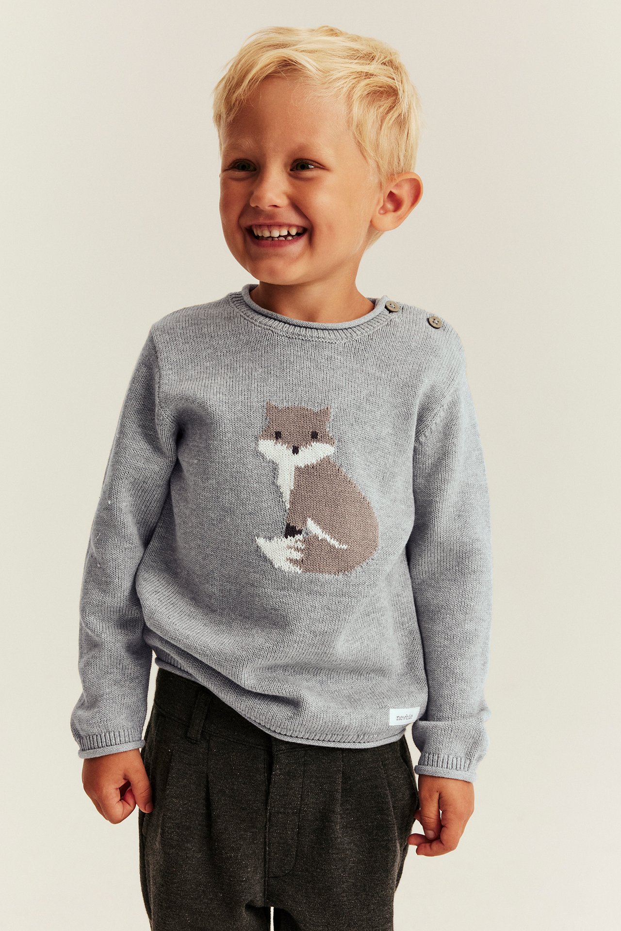 Kind in hellgrauem Fuchs-Pullover mit Schulterknöpfen und dunkelgrauer Hose, Vorderansicht.