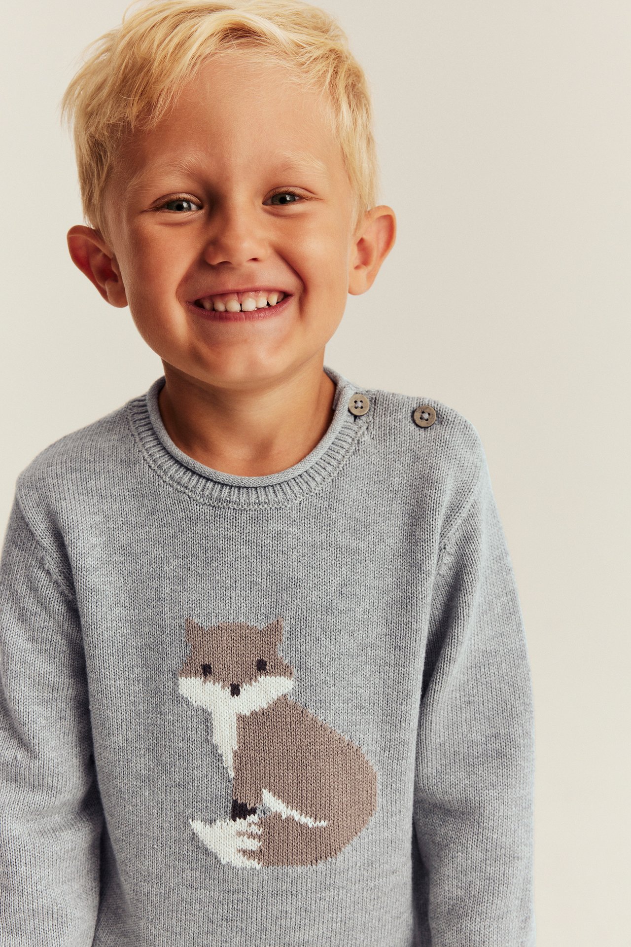 Front view of a kid wearing a light grey knit sweater with a brown and white fox design and shoulder buttons.