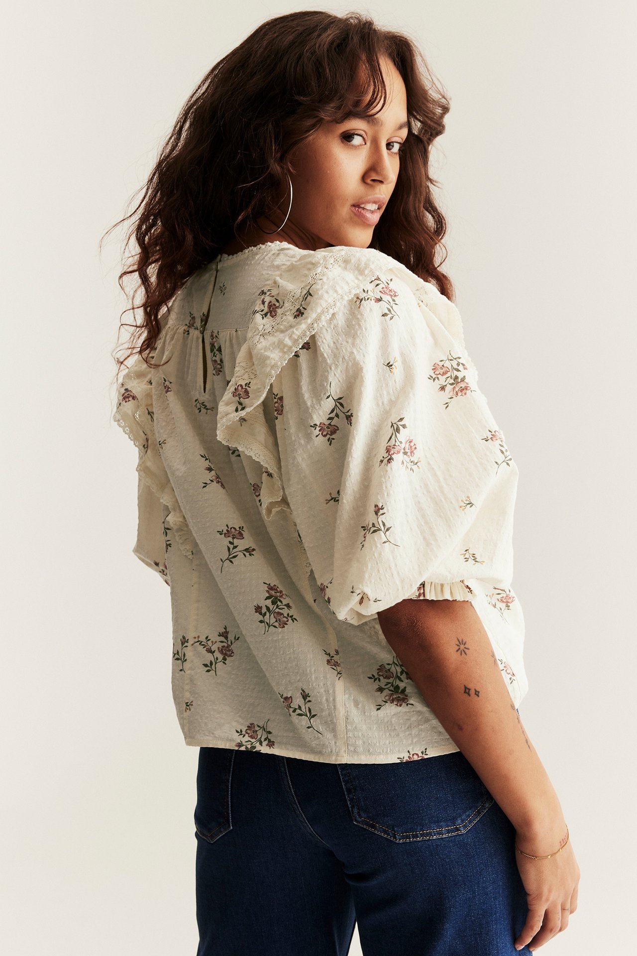 Woman wears off-white floral ruffle blouse with puff sleeves and blue jeans, back-angle view.