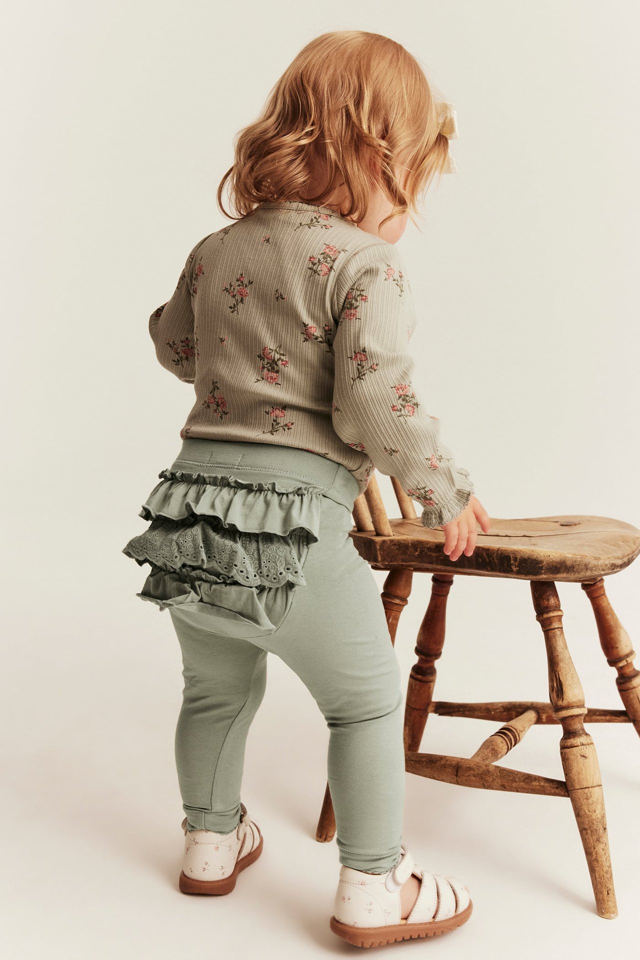 Rückenansicht Baby: grüne Rüschen-Spitzen-Leggings, geblümtes Rippoberteil, weiße Sandalen.