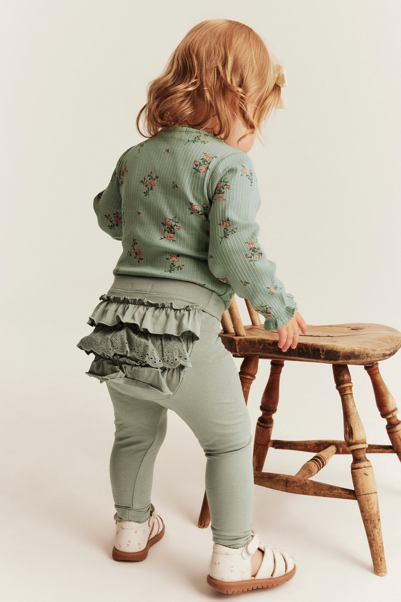 Rückenansicht Baby in salbeigrünem geblümtem Ripptop, Rüschenleggings, weiße Sandalen.