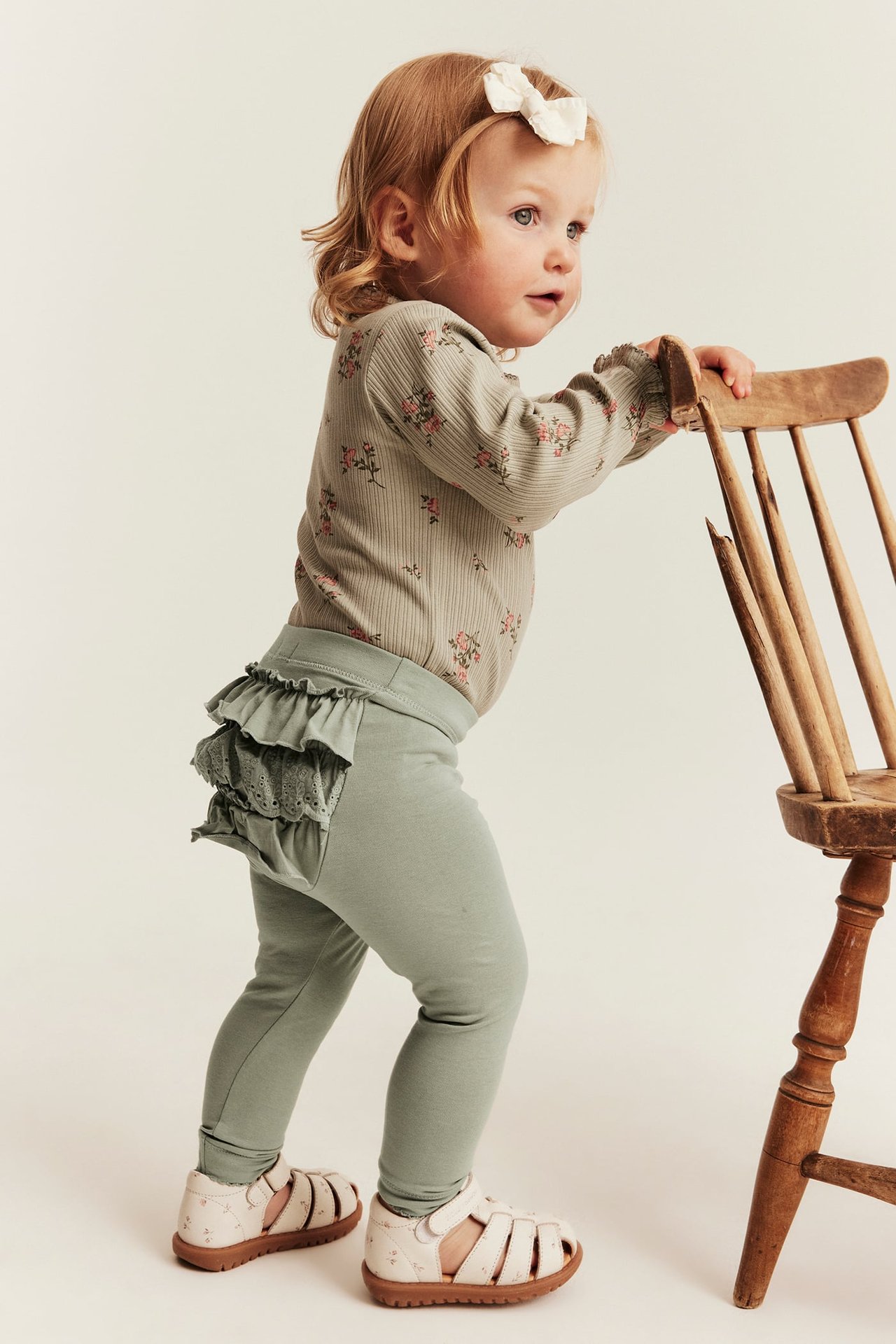 Seitenansicht: Baby-Mädchen trägt grüne Rüschen-Leggings mit Spitze, ein Blumen-Top und weiße Sandalen.