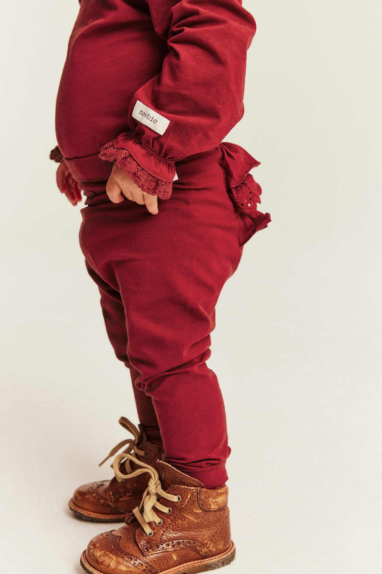 Side view of baby in dark red lace-cuff top, ruffled pants, and brown leather boots.