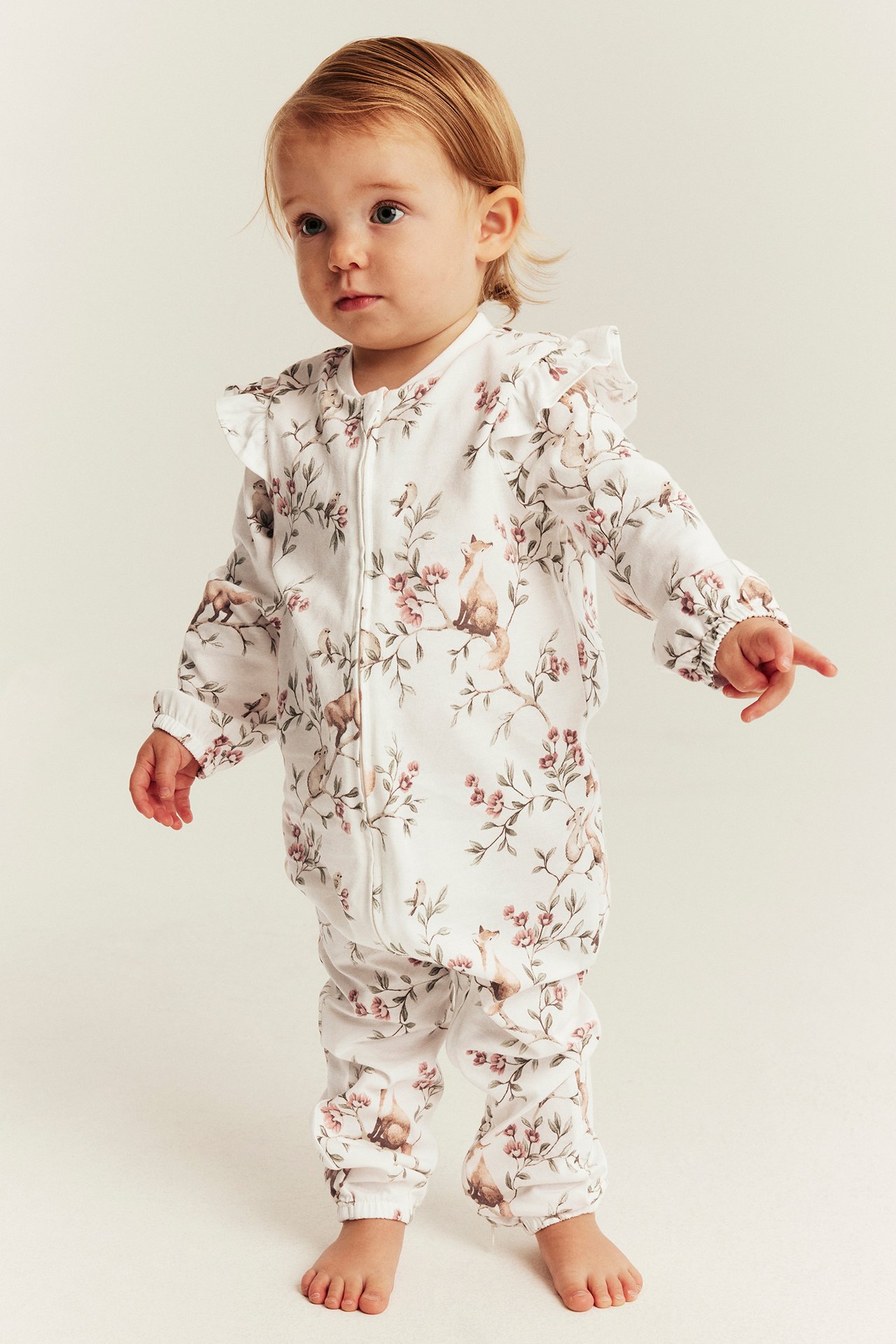 Front view, baby in white long-sleeved romper with floral/animal print, front zipper, ruffles.