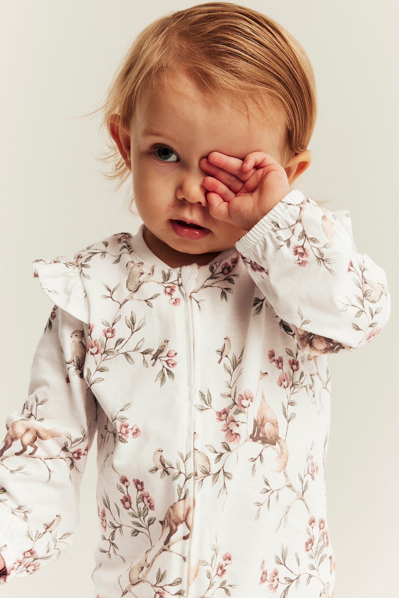 Front view of baby in white long-sleeved sleepsuit with pink floral, animal print, ruffled collar.