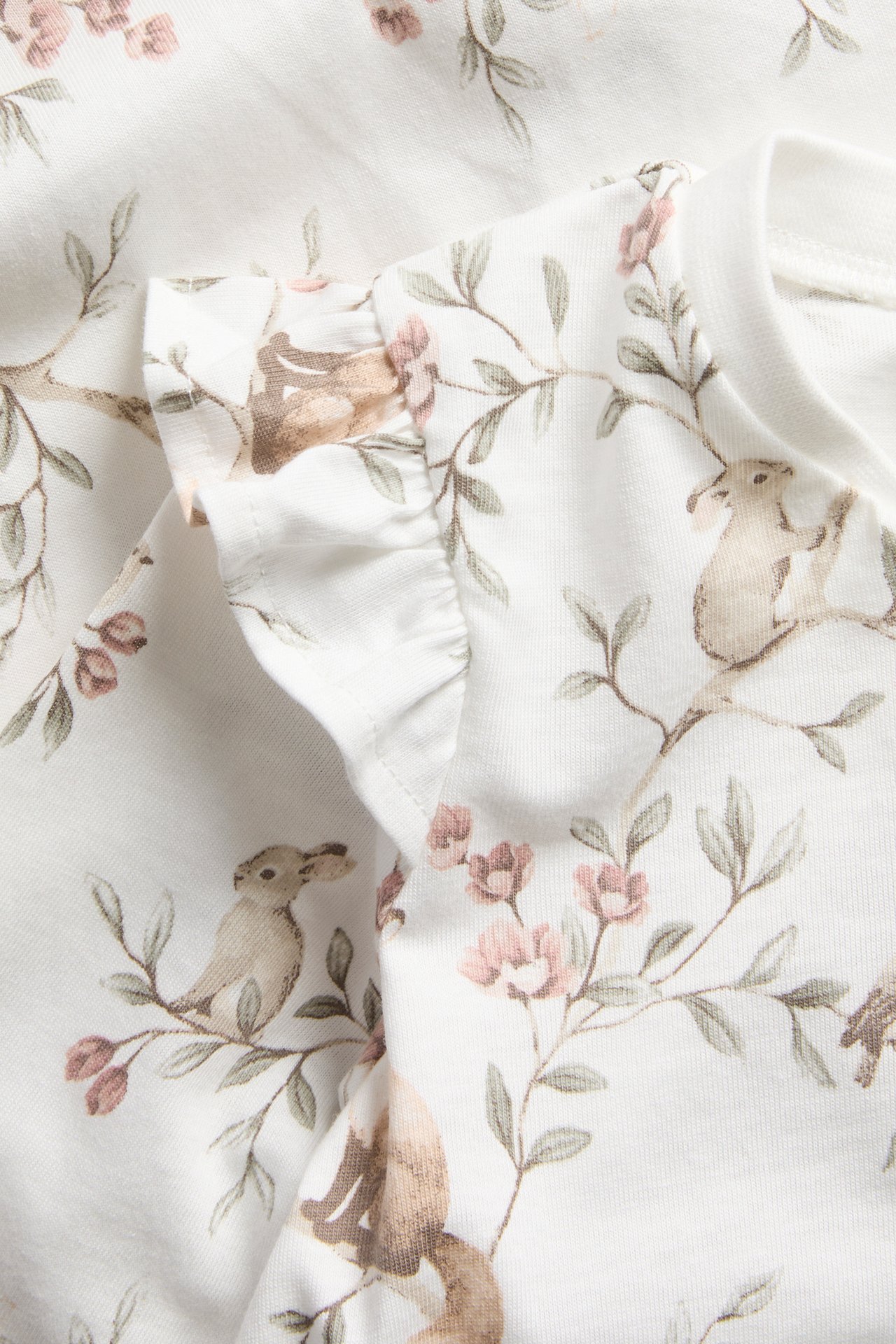 Close-up of off-white cotton baby pyjamas with ruffle shoulders and a forest animal and flower print.
