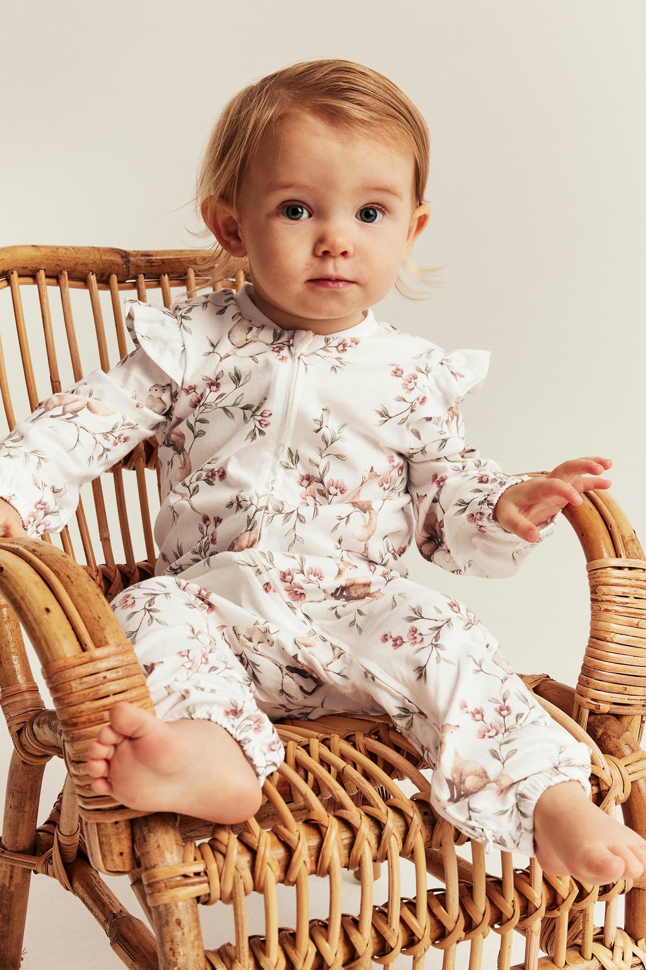 Vorderansicht eines Babys in einem weißen Overall mit Blumen-, Tiermuster, Reißverschluss und Rüschen.
