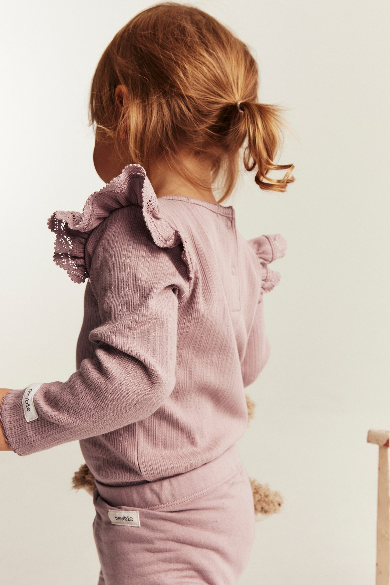 Rückenansicht Kind: Lila geripptes Langarmshirt mit Schulterrüschen und Spitze, plus passende Hose.