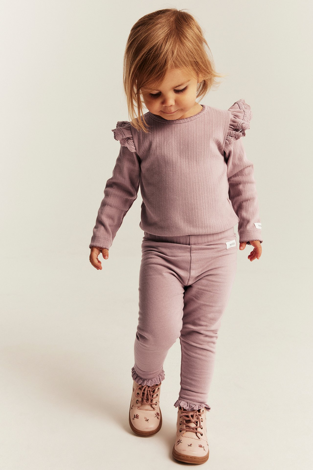 Vorderansicht eines Kindes in lila geripptem Langarm-Set und rosa floralen Stiefeletten.
