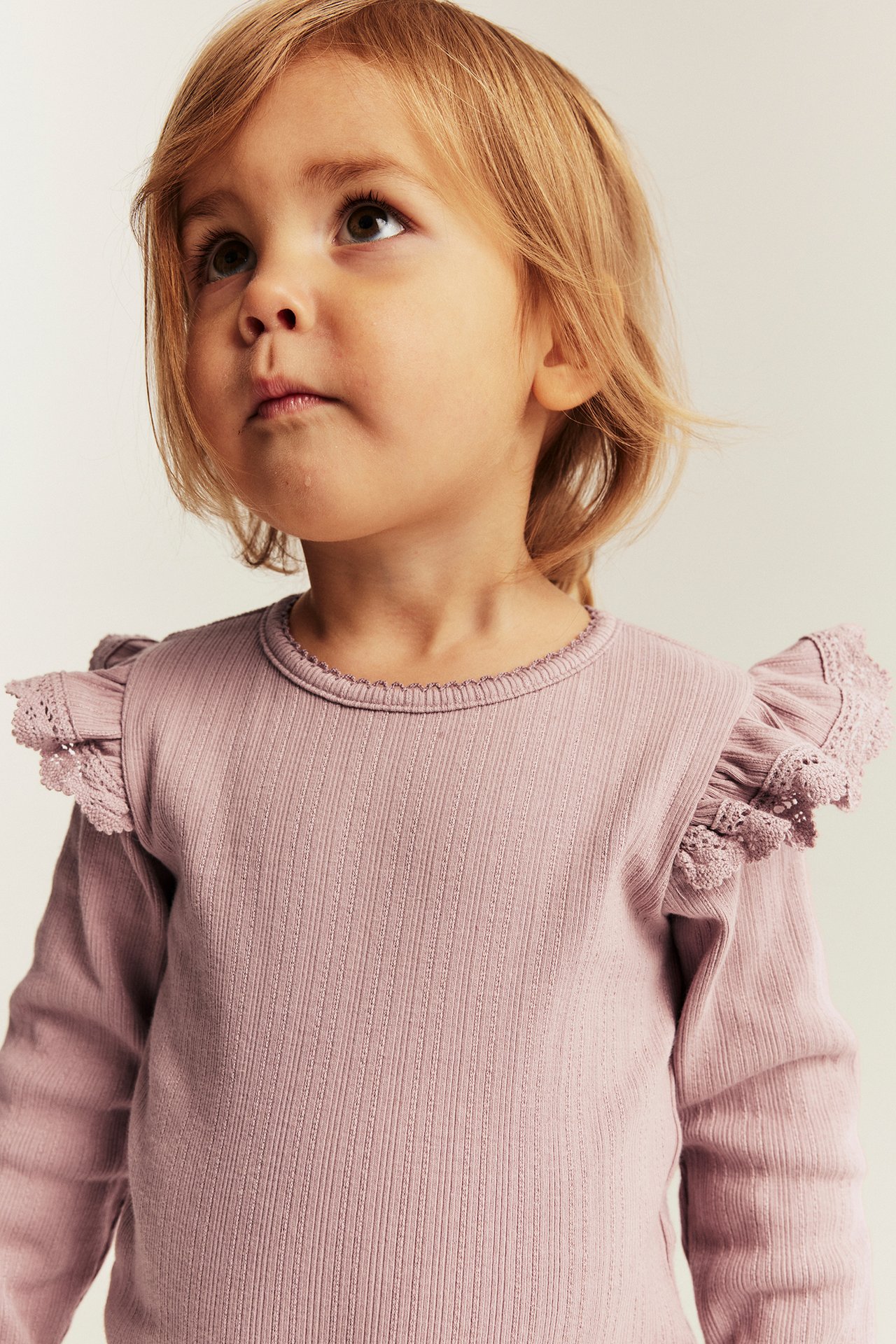 Nahaufnahme eines Kindes, das ein geripptes, langärmeliges, hellviolettes Top mit Spitzenrüschen an den Schultern trägt.