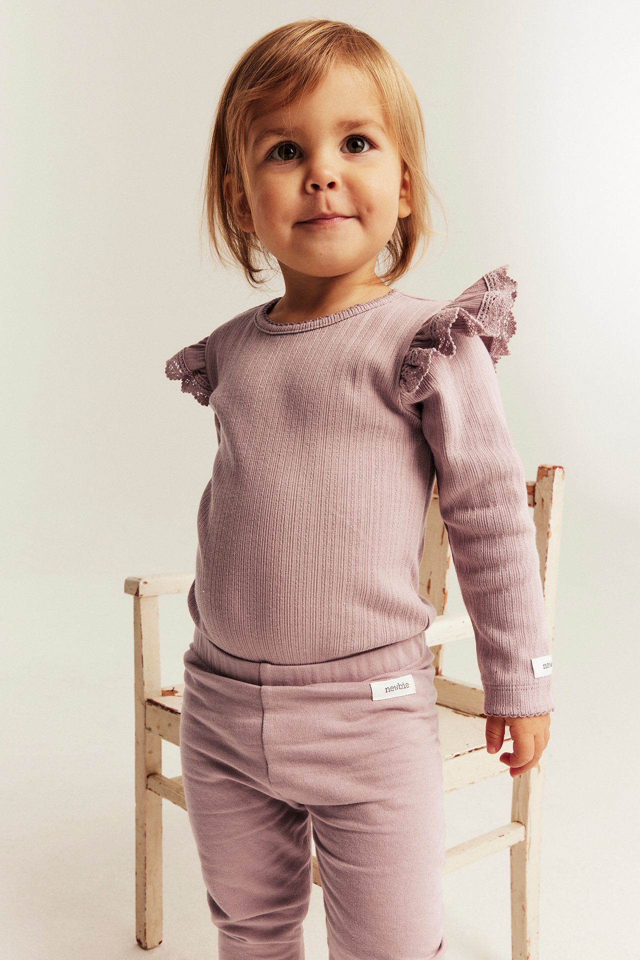 Vorderansicht eines kleinen Mädchens in lila geripptem Langarmshirt mit Rüschen an den Schultern und passenden Leggings.