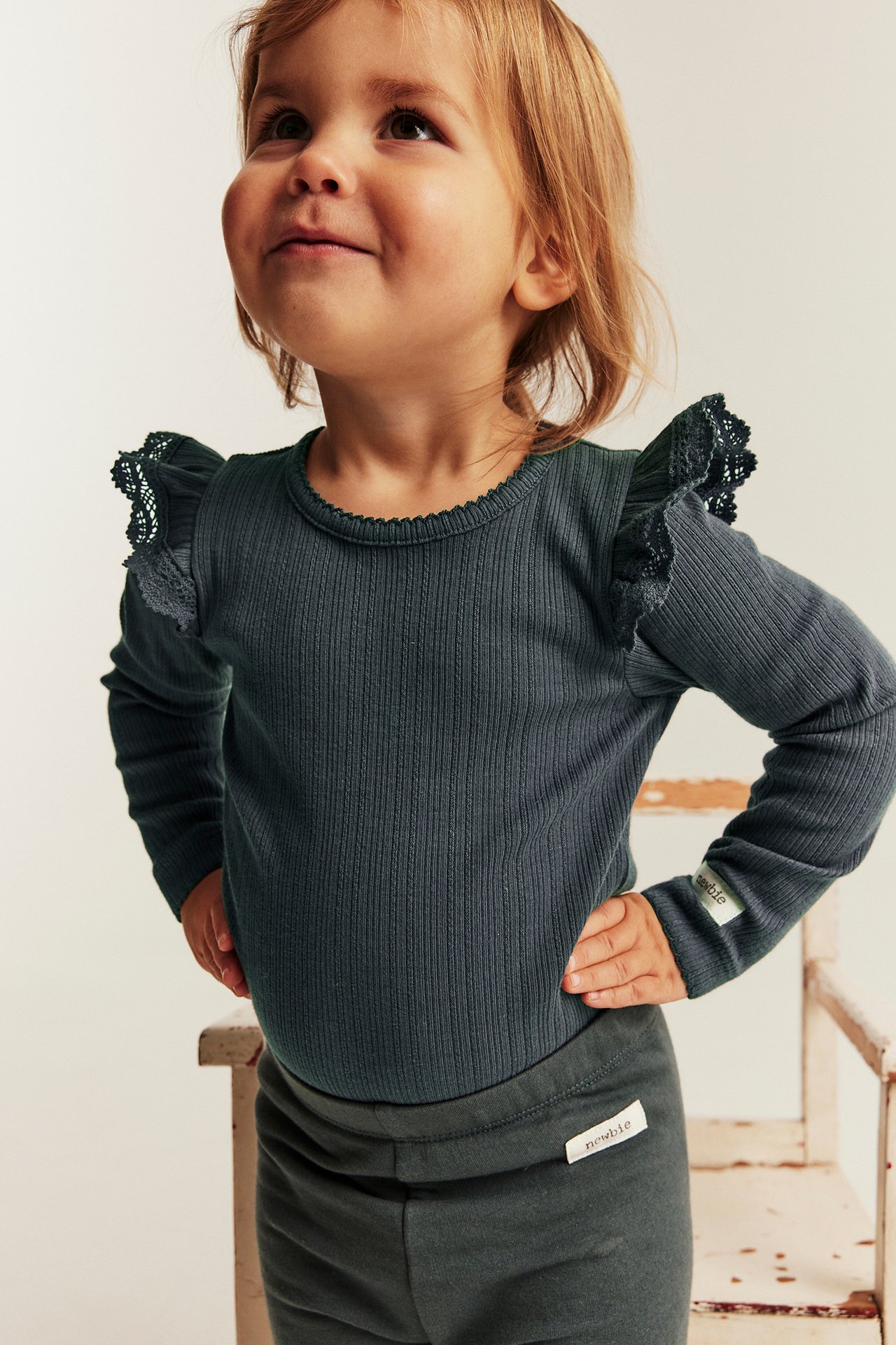 Frontansicht eines Kindes in dunklem Petrol geripptem Langarmshirt mit Spitzen an den Schultern und passenden Leggings.