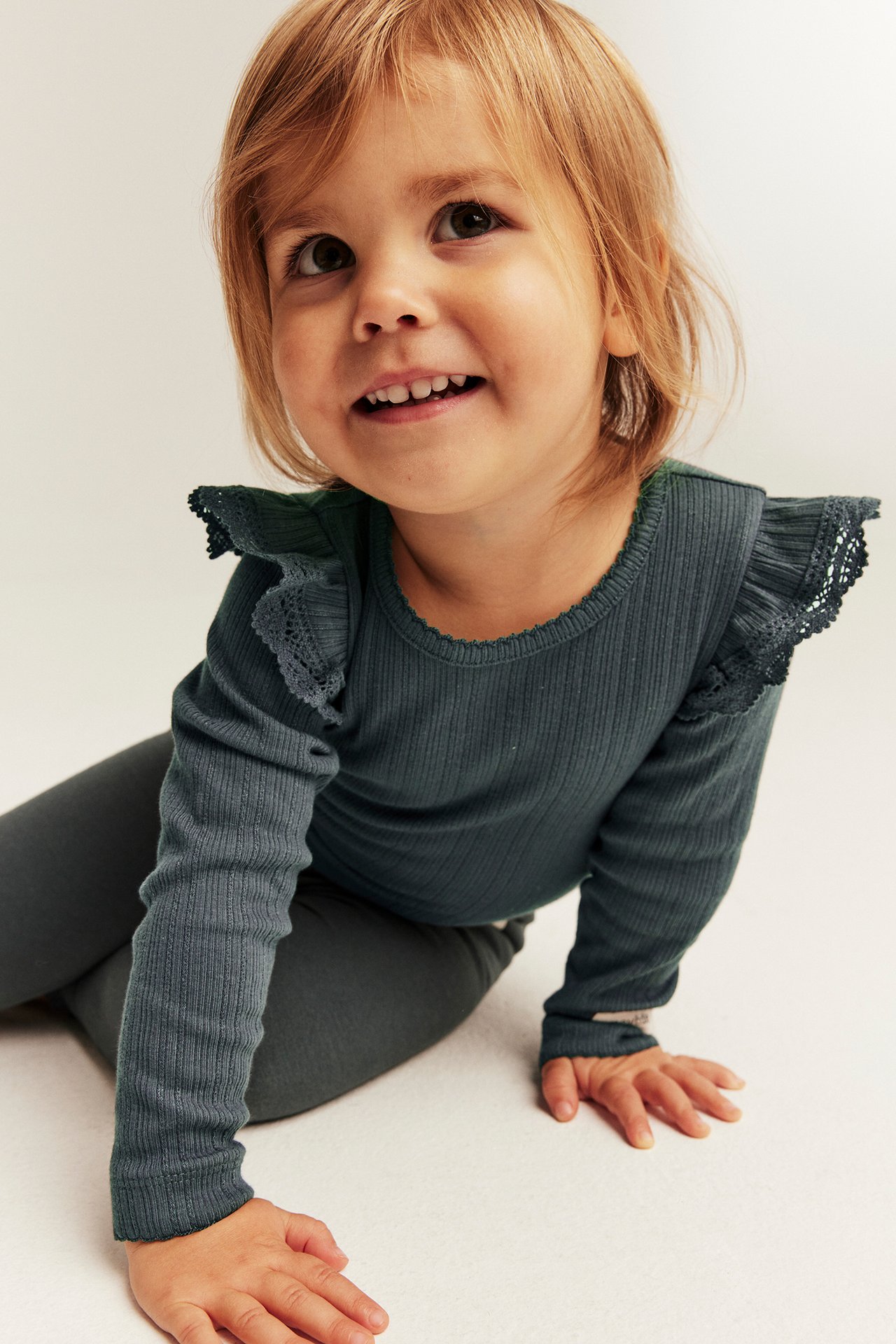 Kind trägt petrolfarbenes geripptes Langarmshirt mit Rüschenspitze und dunkelgraue Leggings.