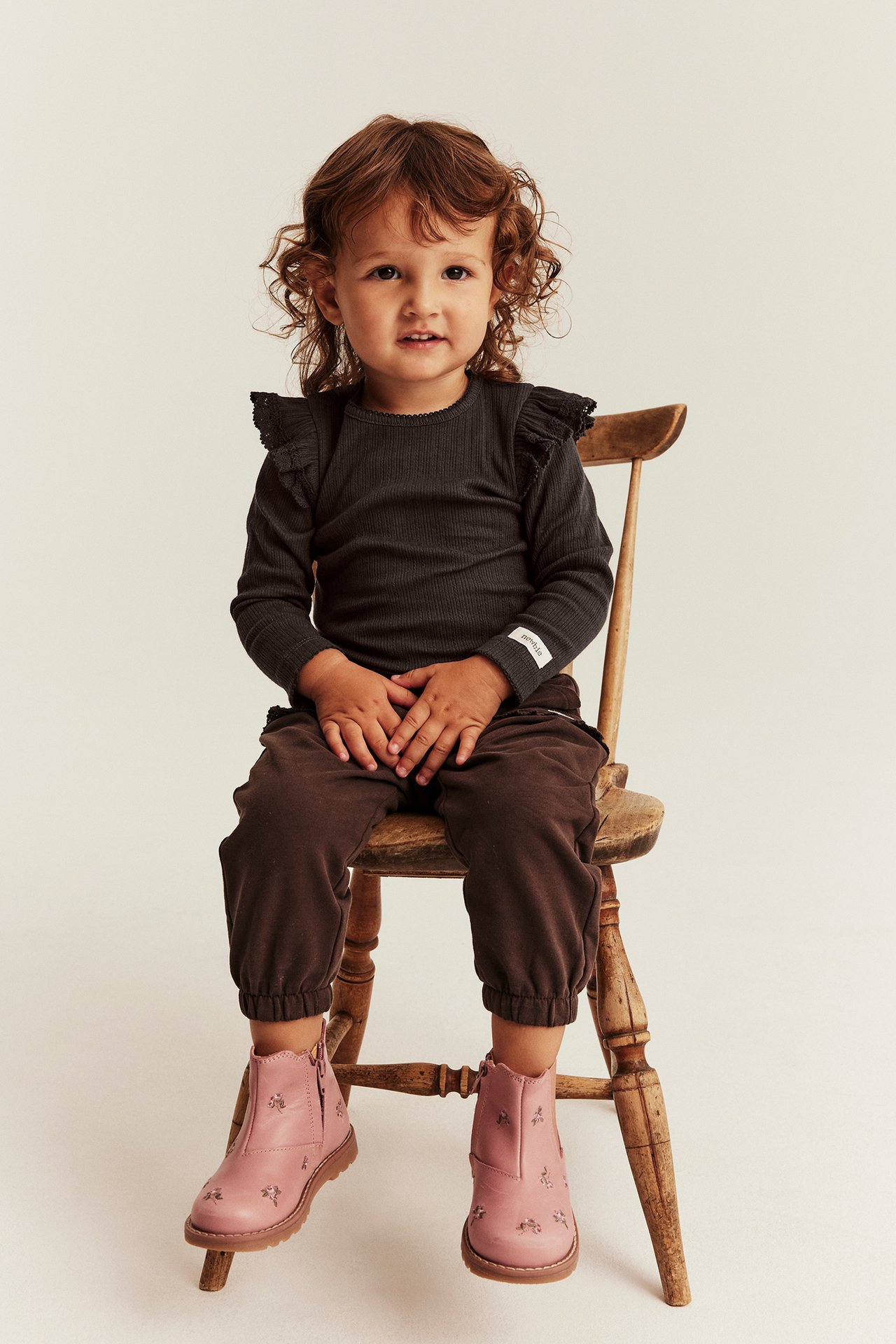 Front view of a kid wearing pink embroidered boots, dark grey ribbed top, and dark brown pants.