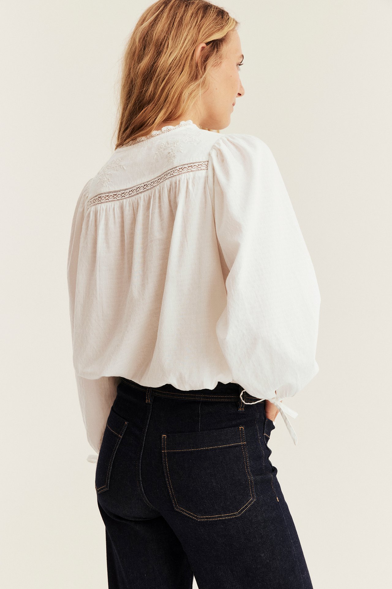 Back view of a female model wearing a white loose-fit embroidered blouse with dark blue jeans.