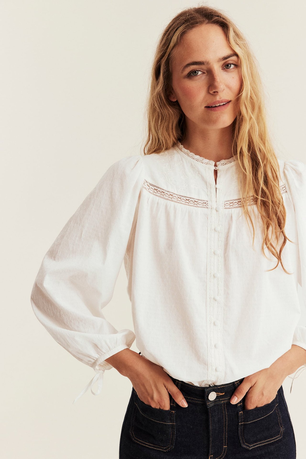 Front view, female model in a white lace-detailed long-sleeve blouse and dark blue jeans.