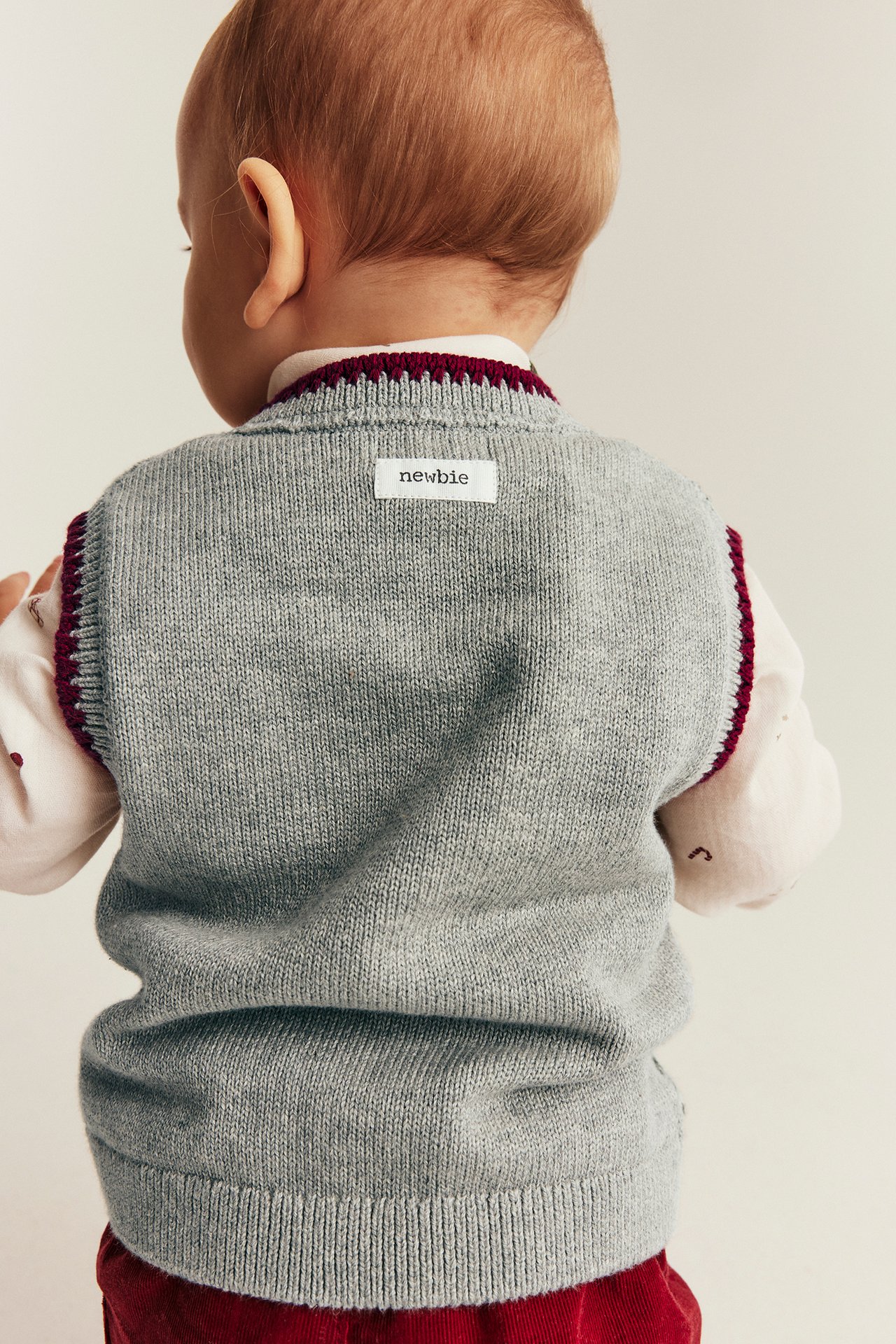 Baby in grauer Strick-V-Weste mit weinrotem Rand über weißem Hemd und roter Hose, Rückenansicht.