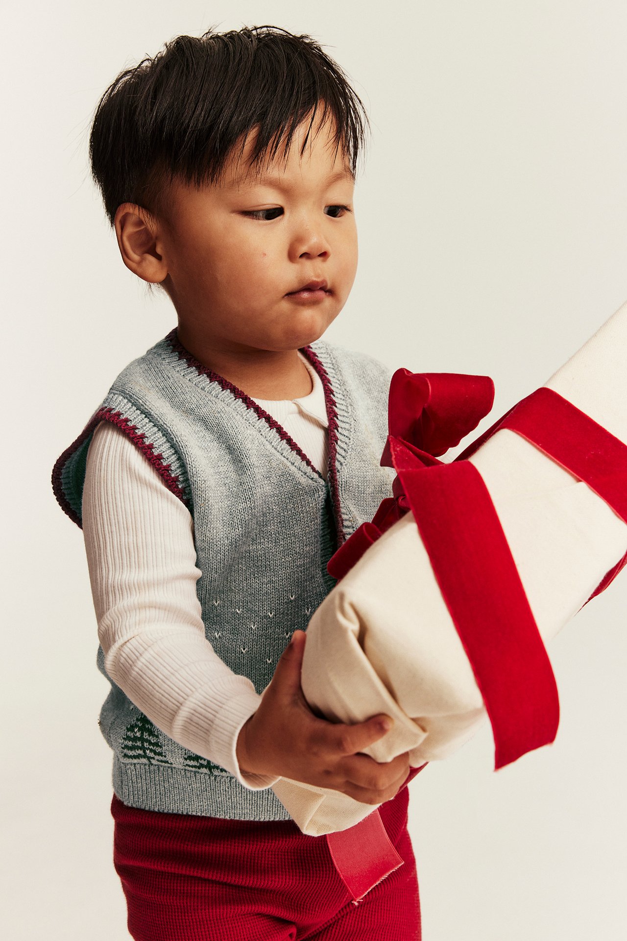 Front view of a kid in a light blue knitted vest, white shirt, and red pants, holding a gift.