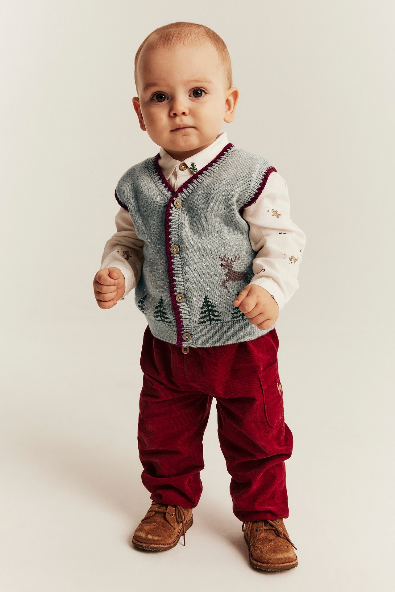 Baby in grey knitted vest with reindeer, printed shirt, red corduroy pants, and brown shoes.