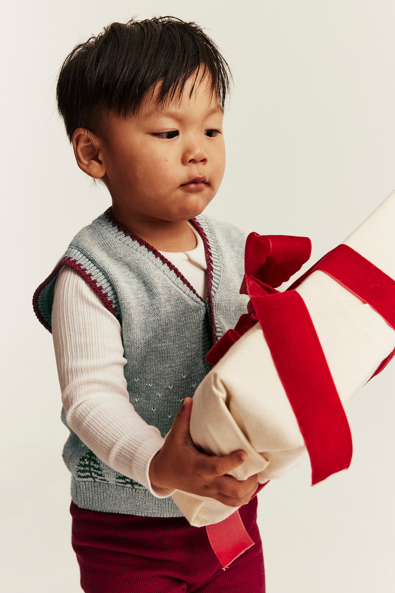 Kind trägt hellblaue Weste mit Bordeaux-Rand, Baummuster, weißes Oberteil, Bordeaux-Hose, hält Geschenk.