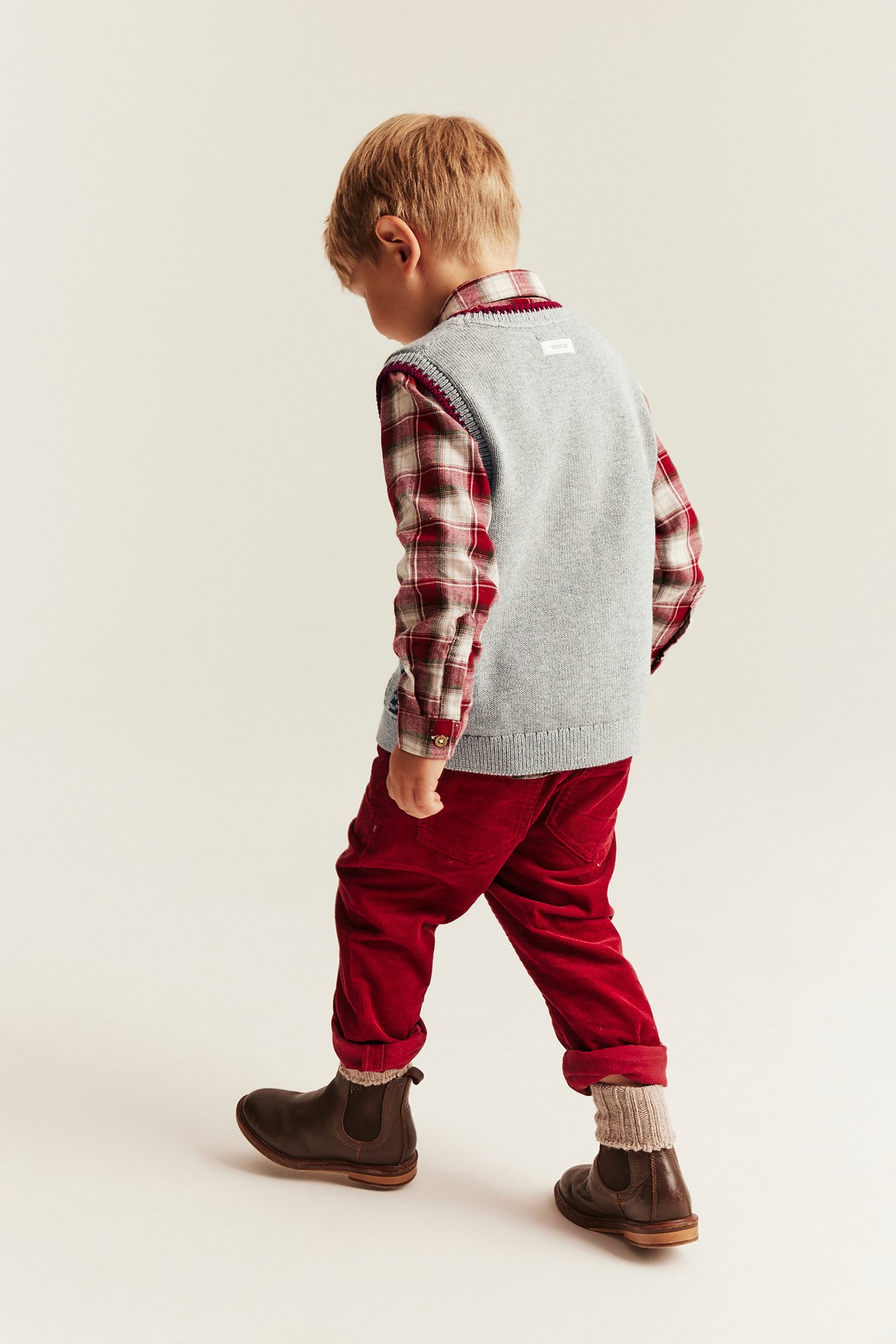 Rückenansicht Kind: graue Strickweste, rot kariertes Hemd, rote Cordhose, braune Stiefel, helle Socken.