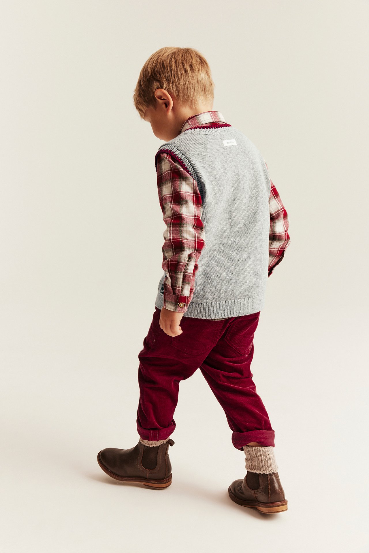 Kinder-Rückenansicht: graue Strickweste über rot kariertem Hemd, rote Cordhose, braune Stiefel, helle Socken.